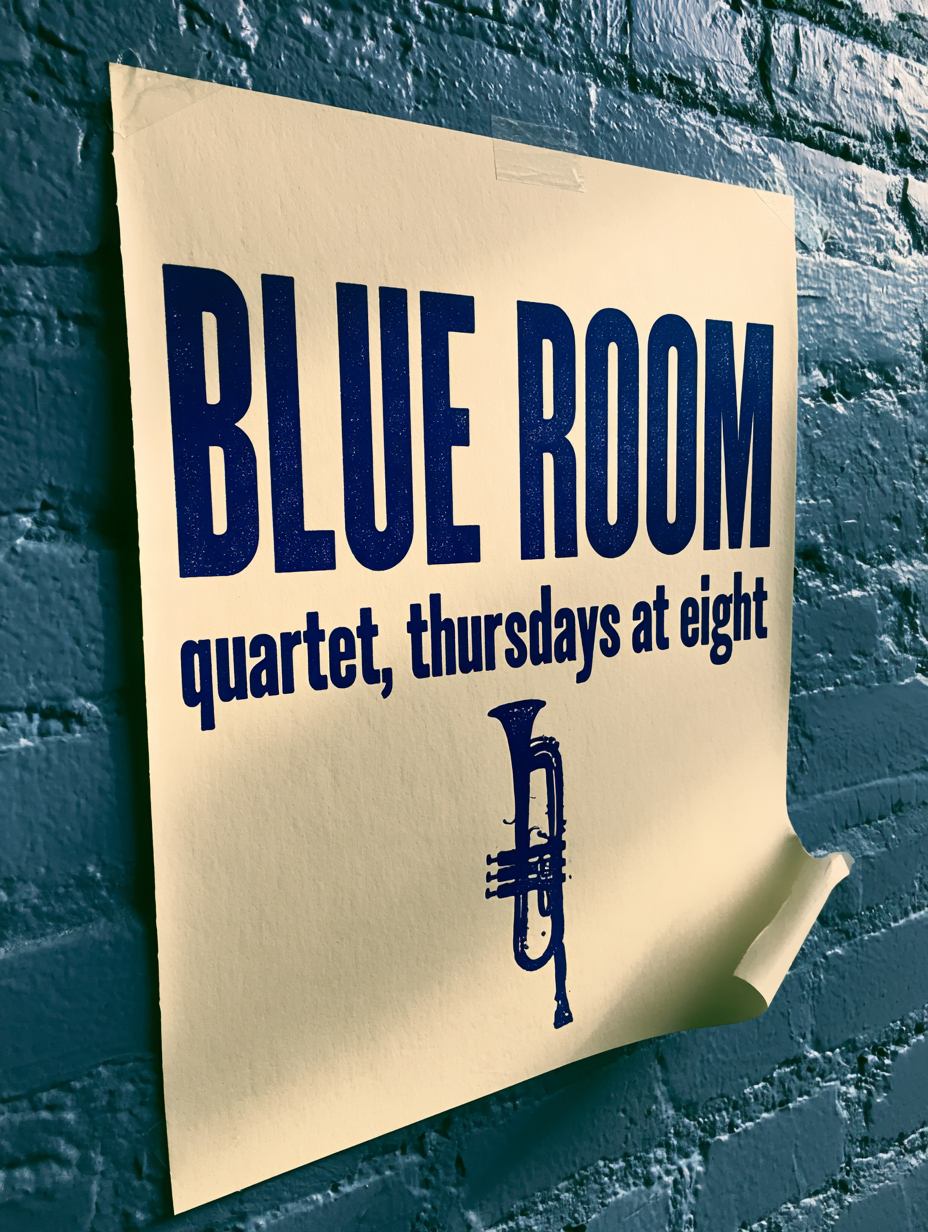Letterpress type, bitten into paper: A letterpress gig poster taped to a painted brick wall, deep indigo ink on heavy cream stock, large headline "BLUE ROOM" with a smaller line "quartet, thursdays at eight" beneath it, a small woodcut trumpet motif, visible plate impression and ink bite in the paper, one corner curling away from the wall, raking evening light across the brick --style raw --s 25 --no watermark, extra text