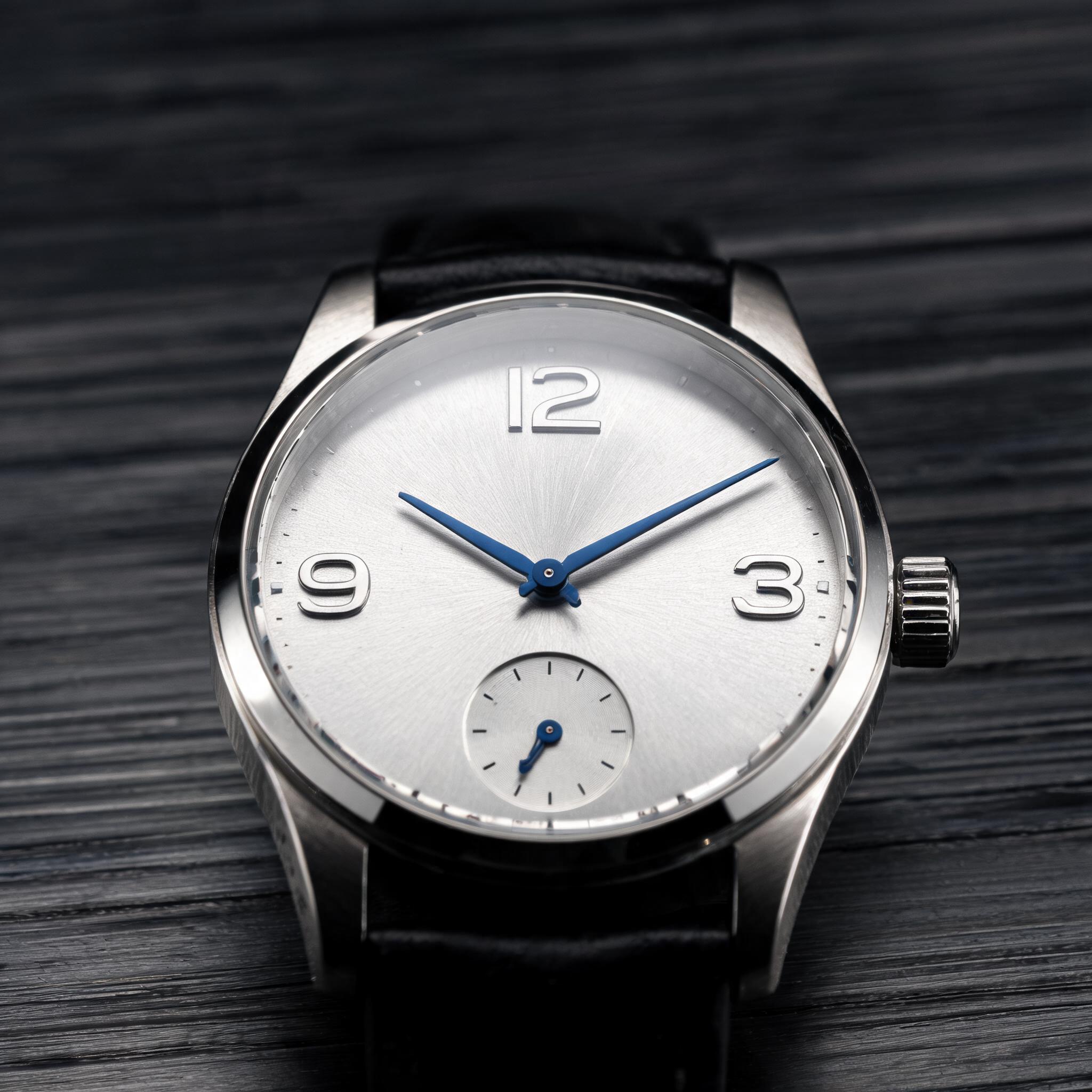 Distinct finishes in one shot: Macro product photograph of a mechanical wristwatch lying on dark brushed steel, square format: a silver sunburst dial with only three large applied numerals, 12 at the top, 3 on the right and 9 on the left, and a small seconds subdial sitting at the six o'clock position in place of a 6, no other numerals anywhere. Blued steel hands, a brushed steel case with polished bevels, a black leather strap curling out of focus. One hard key light from the upper left throwing crisp highlights on the bevels and a soft reflection on the crystal. Photoreal, 100mm macro, shallow depth of field. No text on the dial, no brand name, no logos, no watermark.
